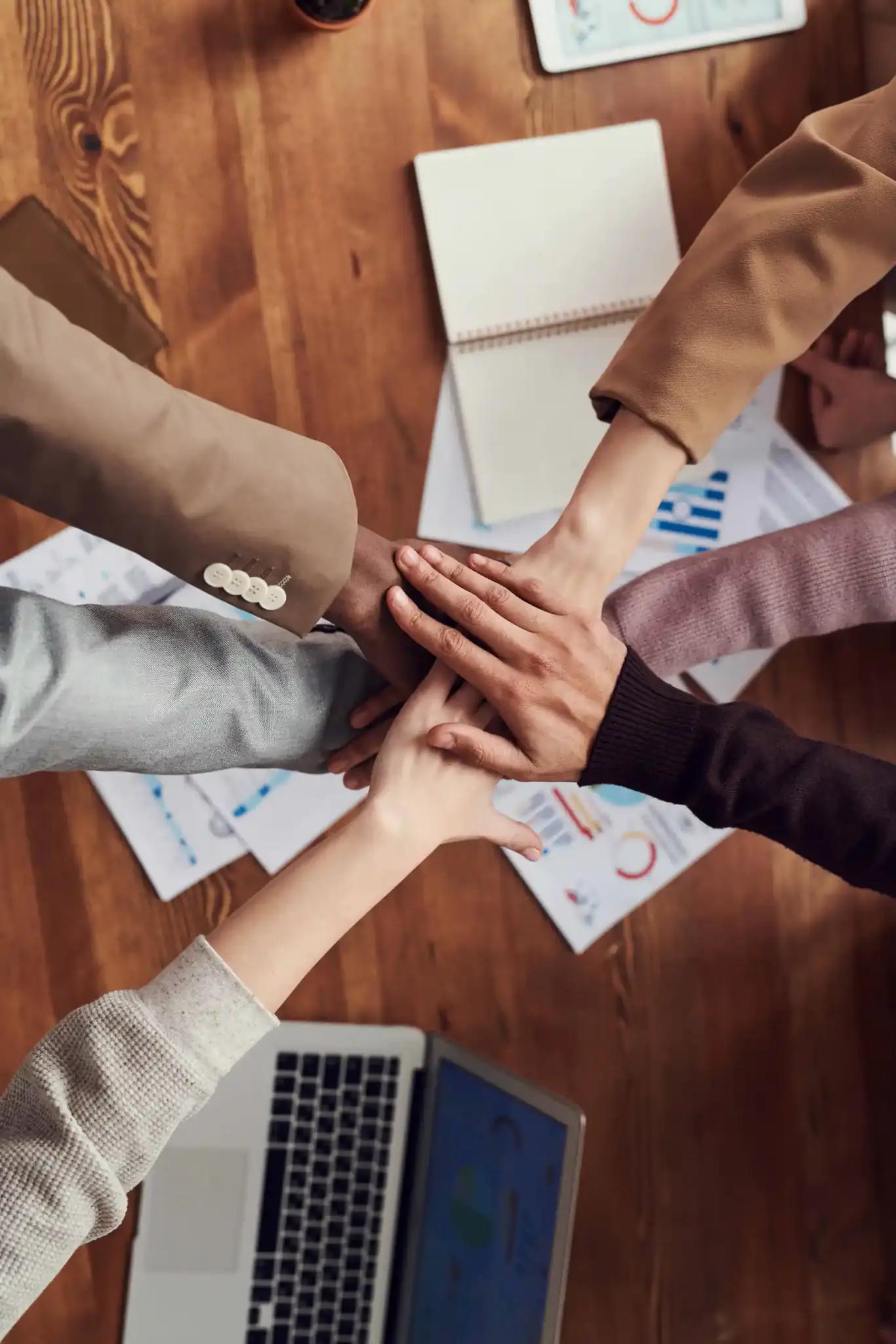 Teammitglieder legen ihre Hände zusammen über einem Tisch mit Laptop und Diagrammen.