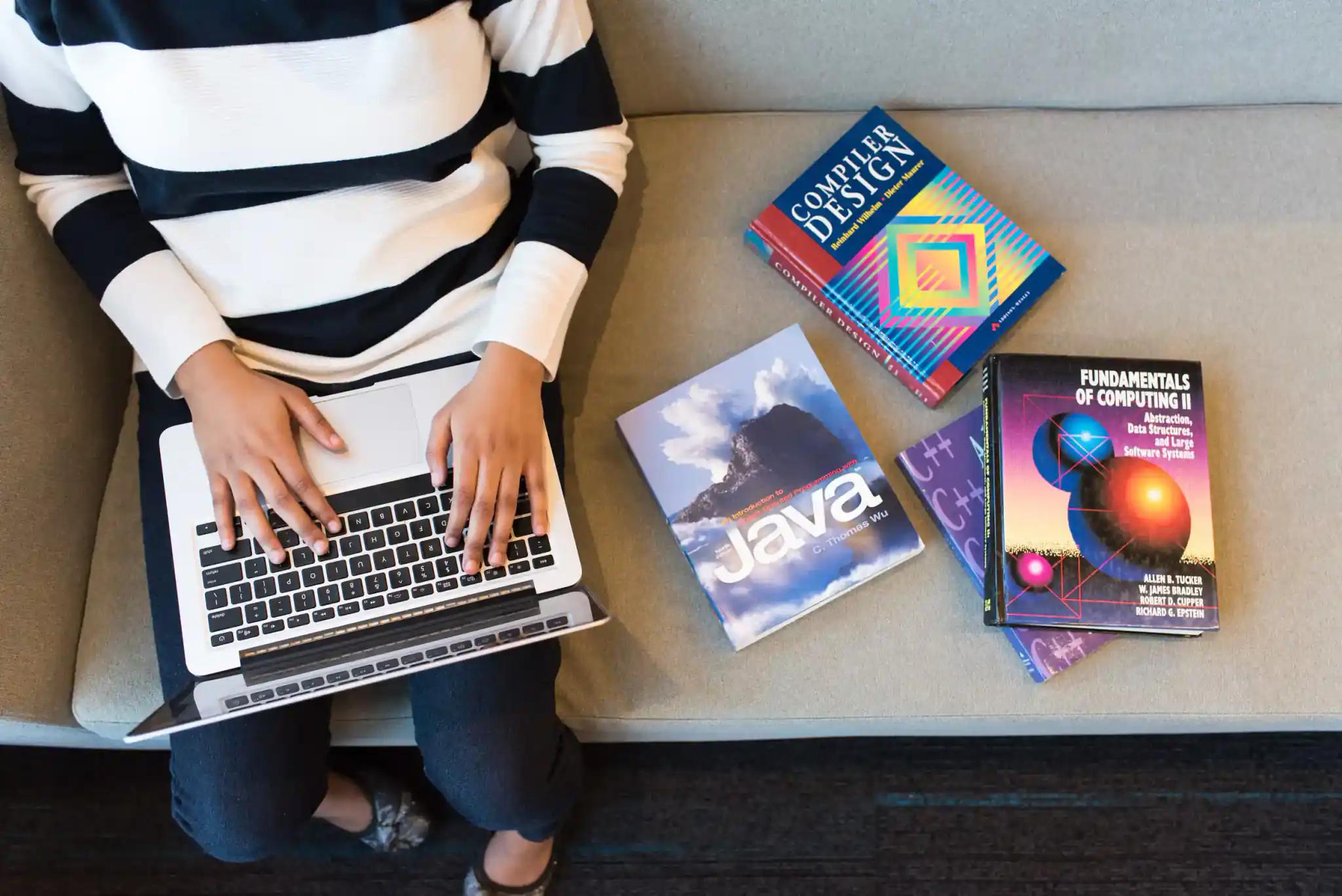 Person tippt auf einem Laptop mit Programmierbüchern neben sich