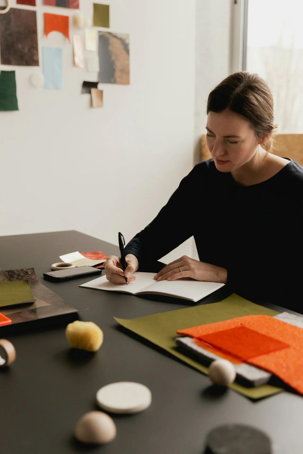 Woman writing in notebook at table with design materials.