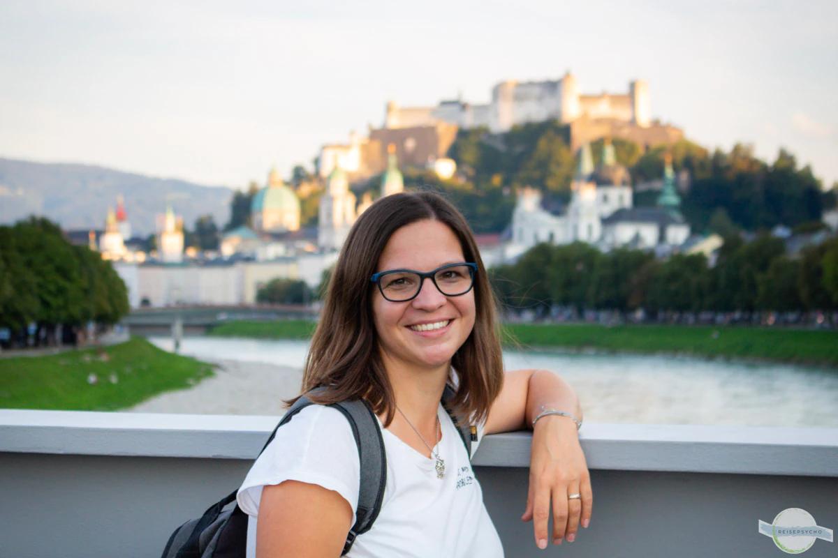 Lächelnde Frau mit Brille vor einer historischen Stadt mit einem Fluss.