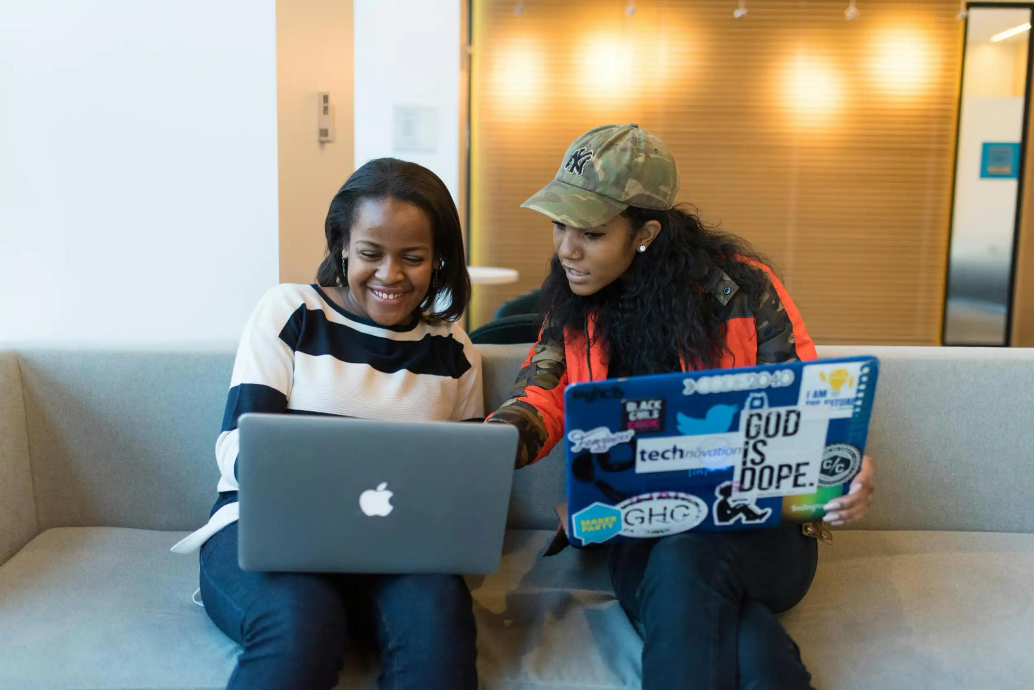 Two women working together on laptops while sitting on a couch.