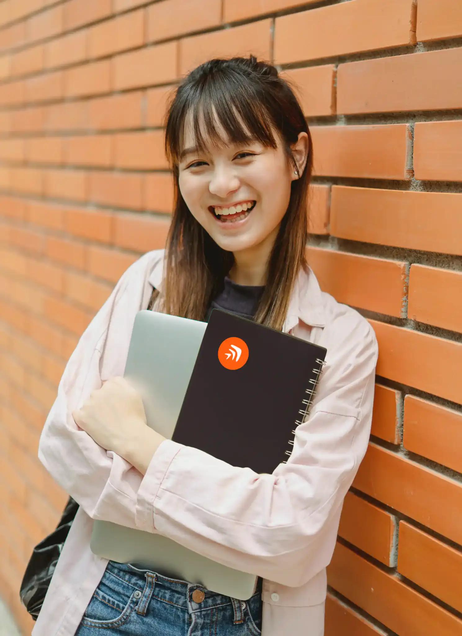 Lächelnde Frau mit Laptop und Notizbuch vor einer Ziegelwand.