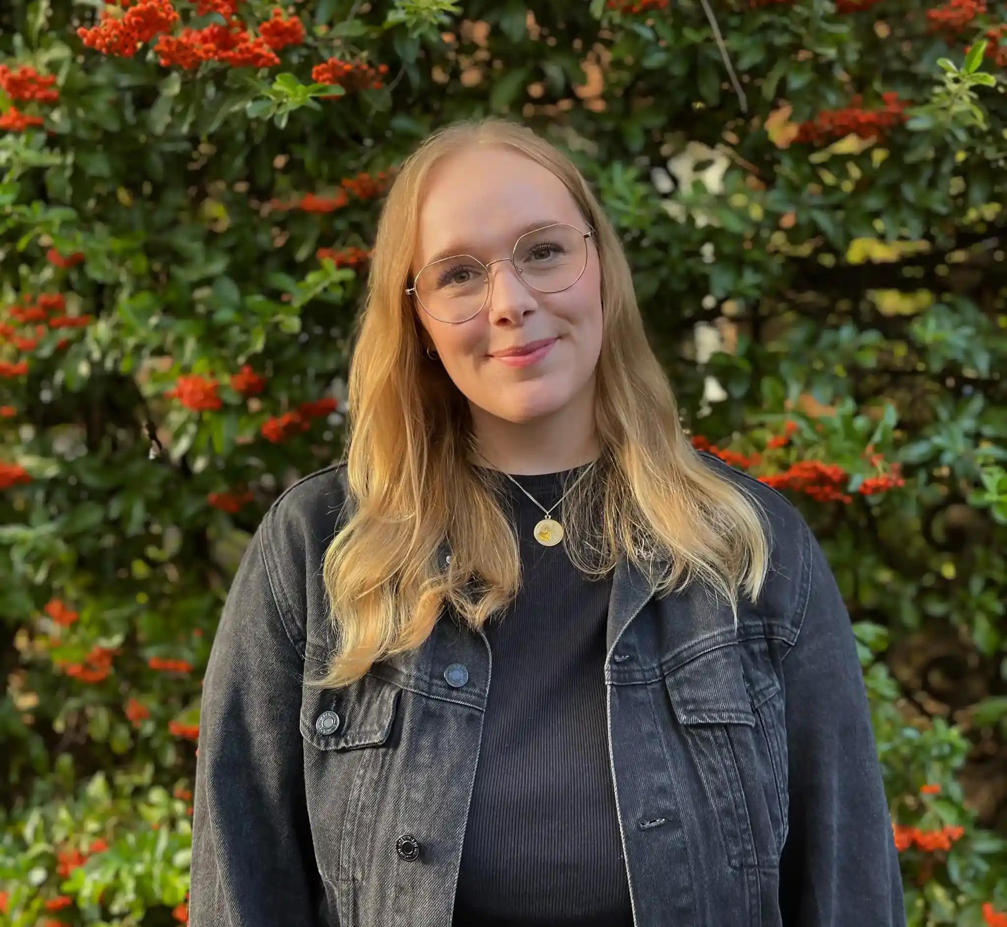 Portrait of a young woman with glasses in front of a plant wall with red berries.