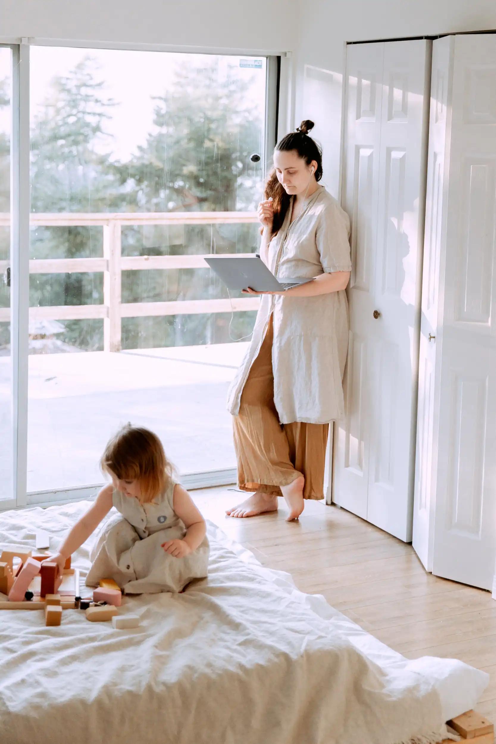 Woman standing with laptop while child plays with wooden toys on bed.