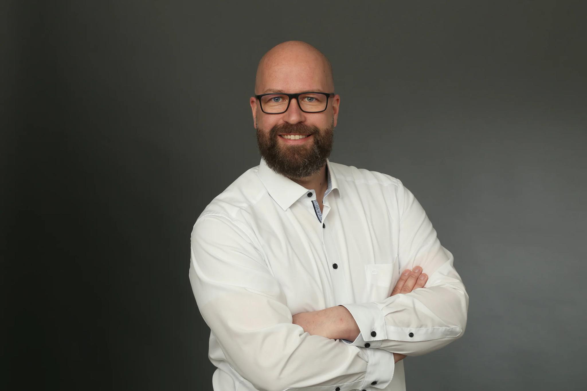 Porträt eines lächelnden Mannes mit Brille und weißem Hemd vor grauem Hintergrund