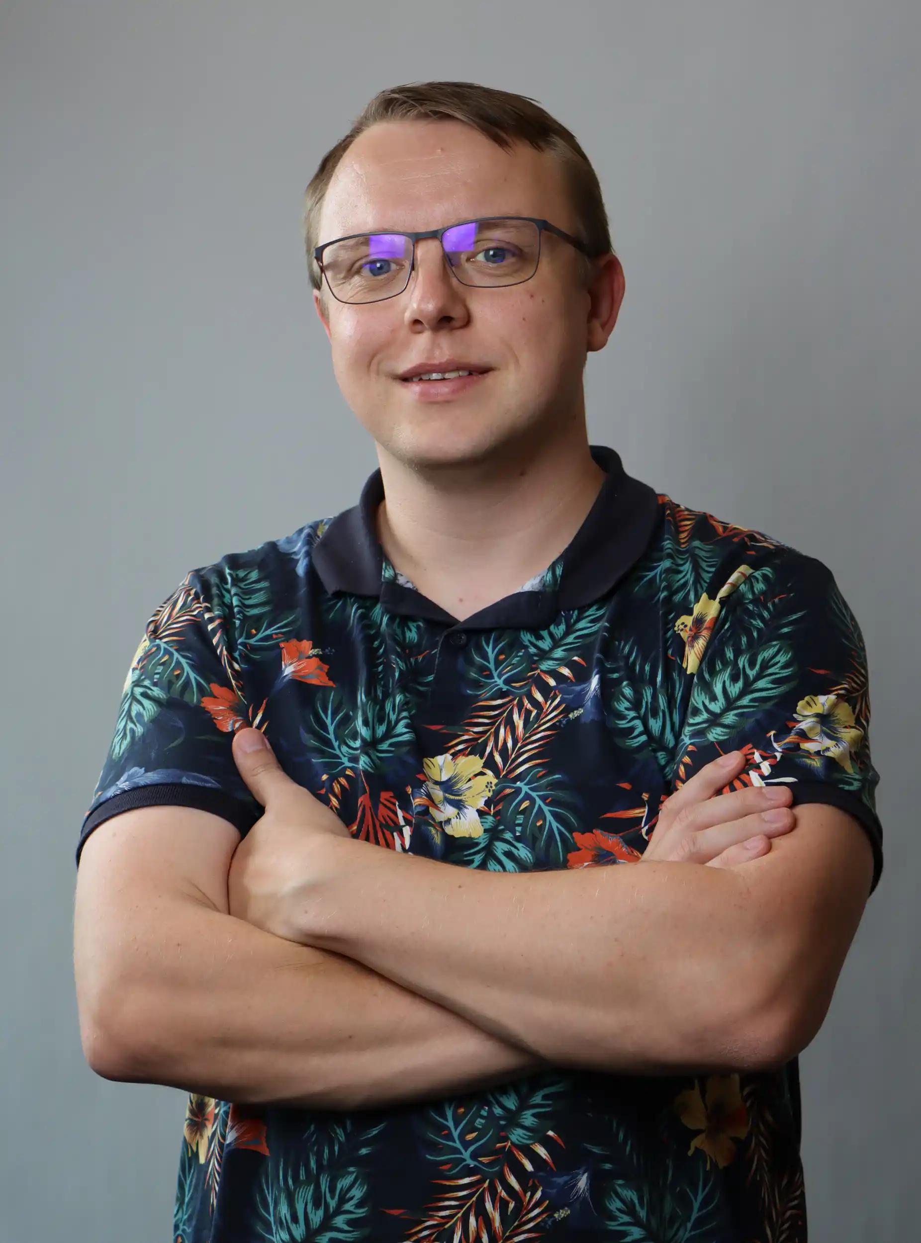Man with glasses and floral shirt with crossed arms against gray background.
