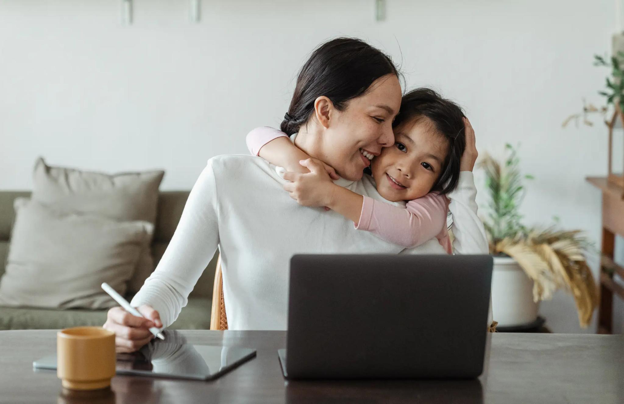 Mother being hugged by daughter while working on laptop.