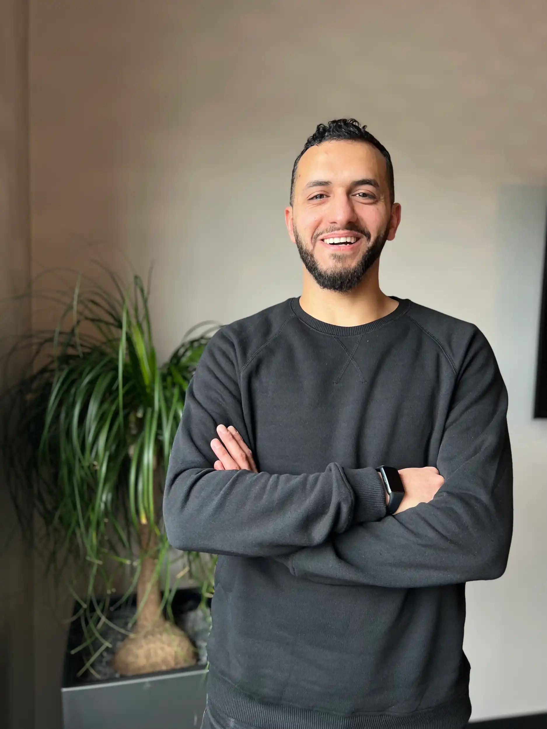 Lächelnder Mann mit verschränkten Armen vor einer Pflanze.