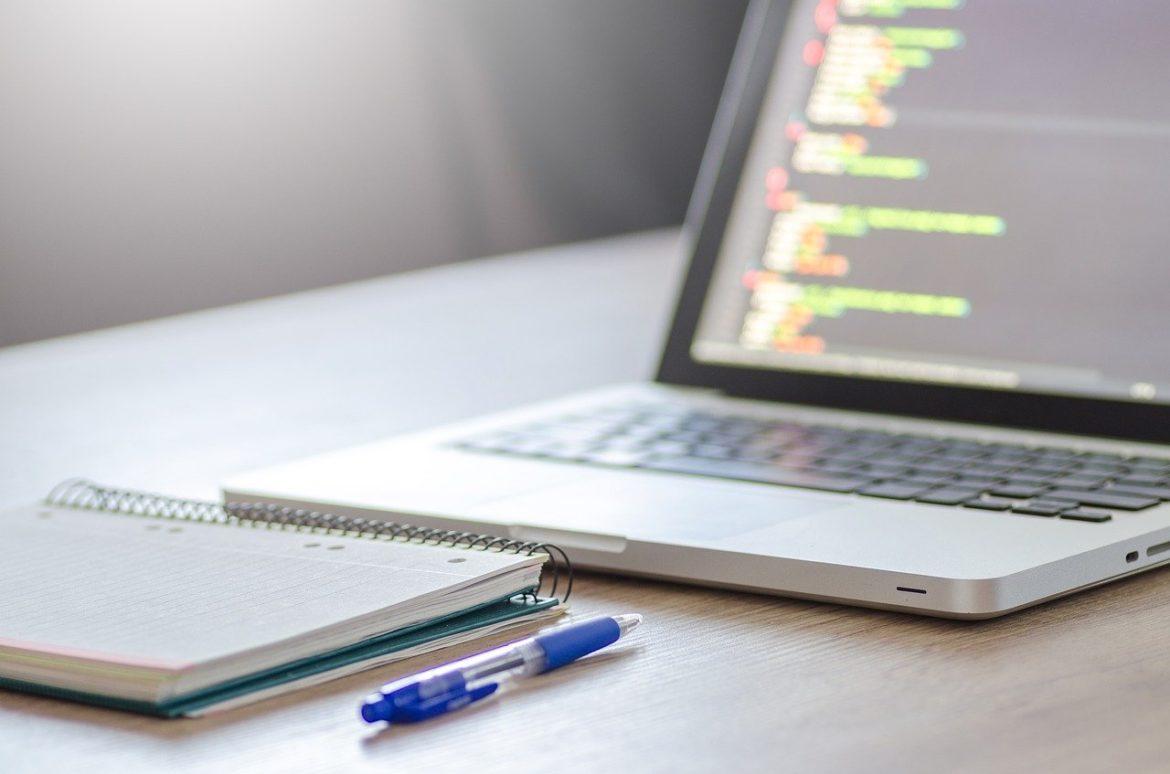 Laptop with programming code, notebook, and pen on desk