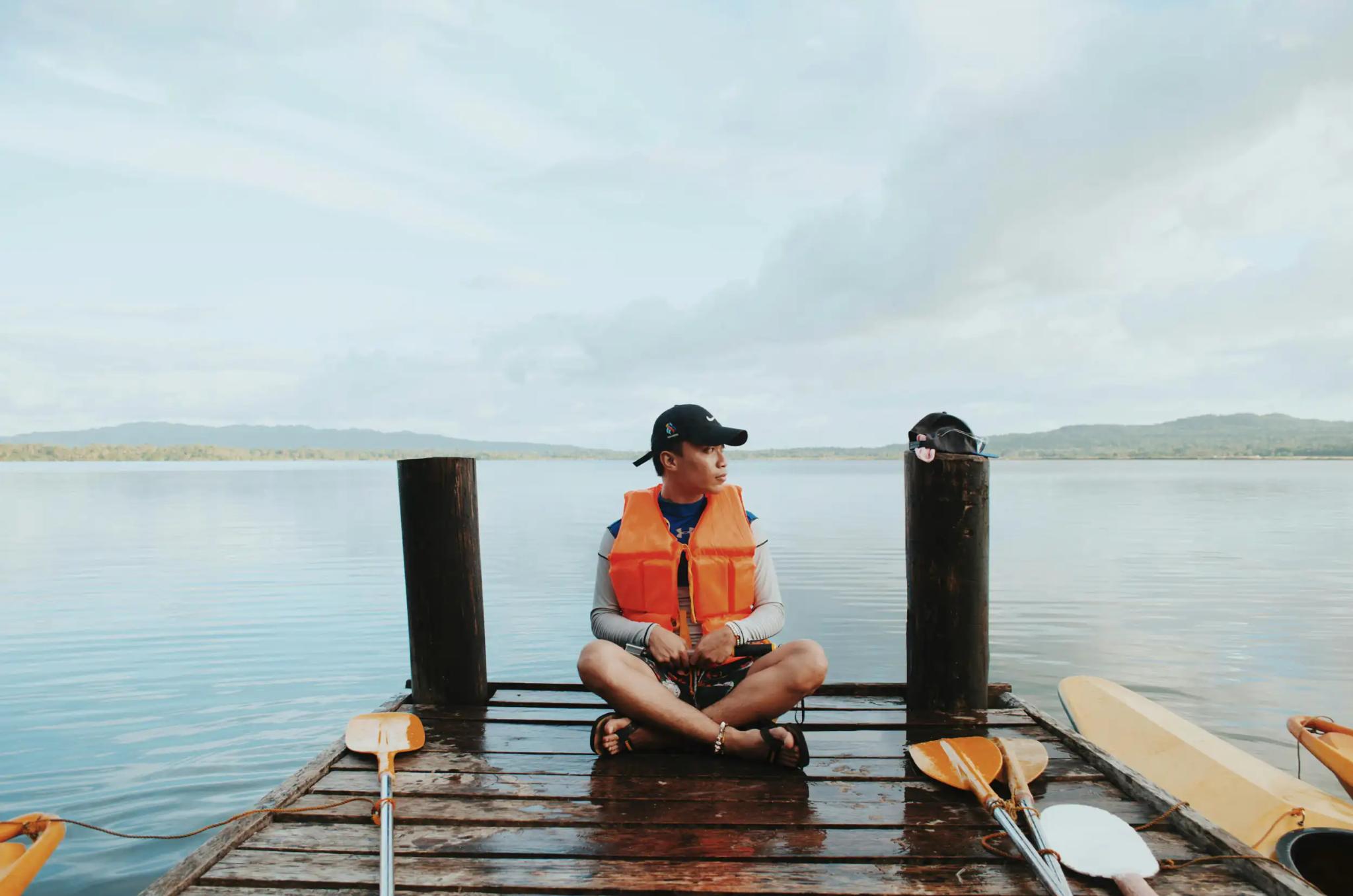 Man wearing life vest sitting on a dock by the water.