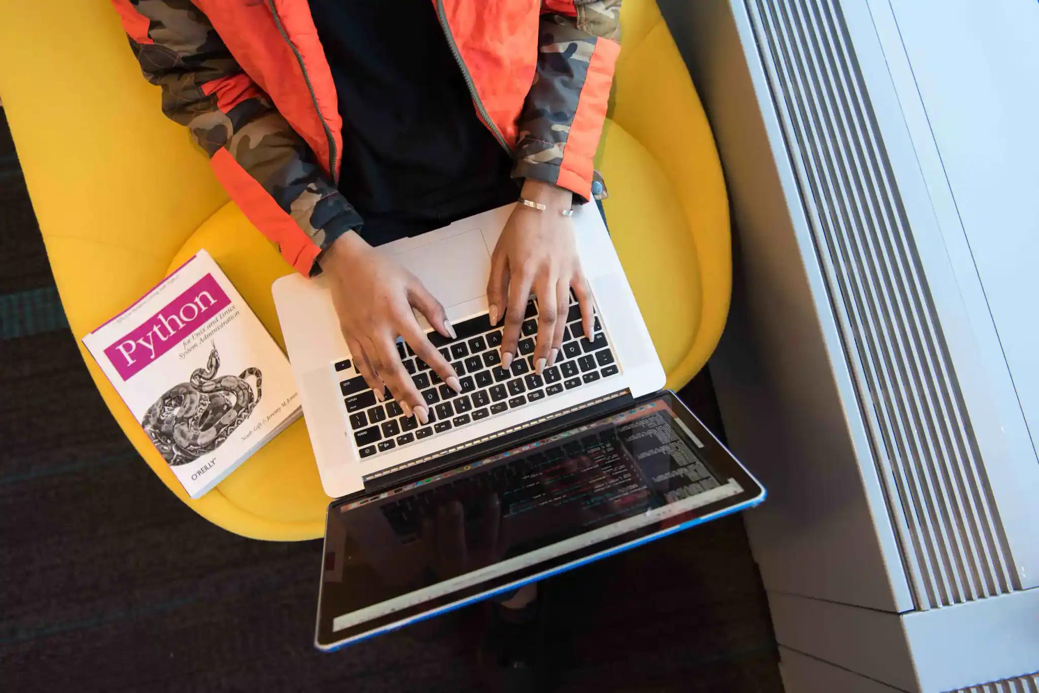 Person typing on laptop next to Python book.