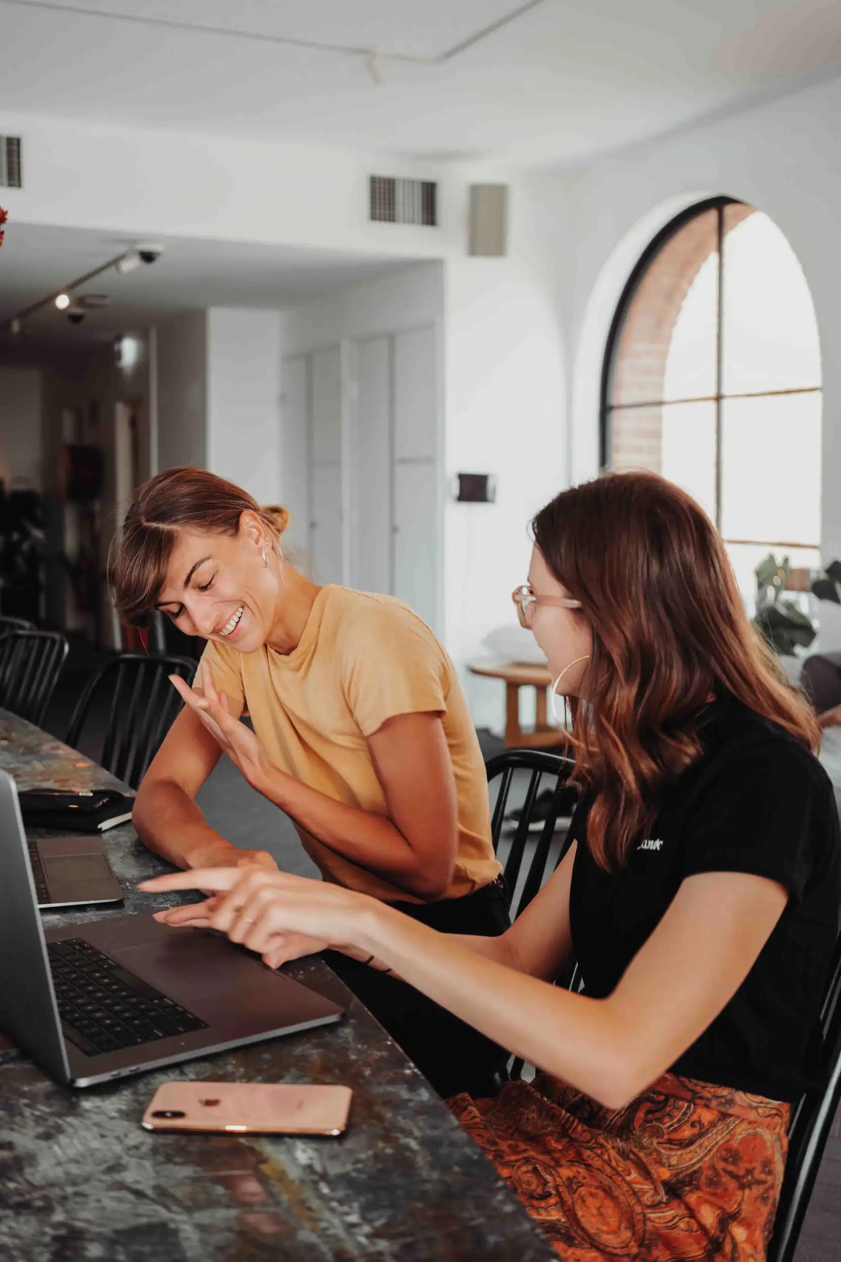 Zwei Frauen lachen und arbeiten gemeinsam an einem Laptop in einem modernen Büro.