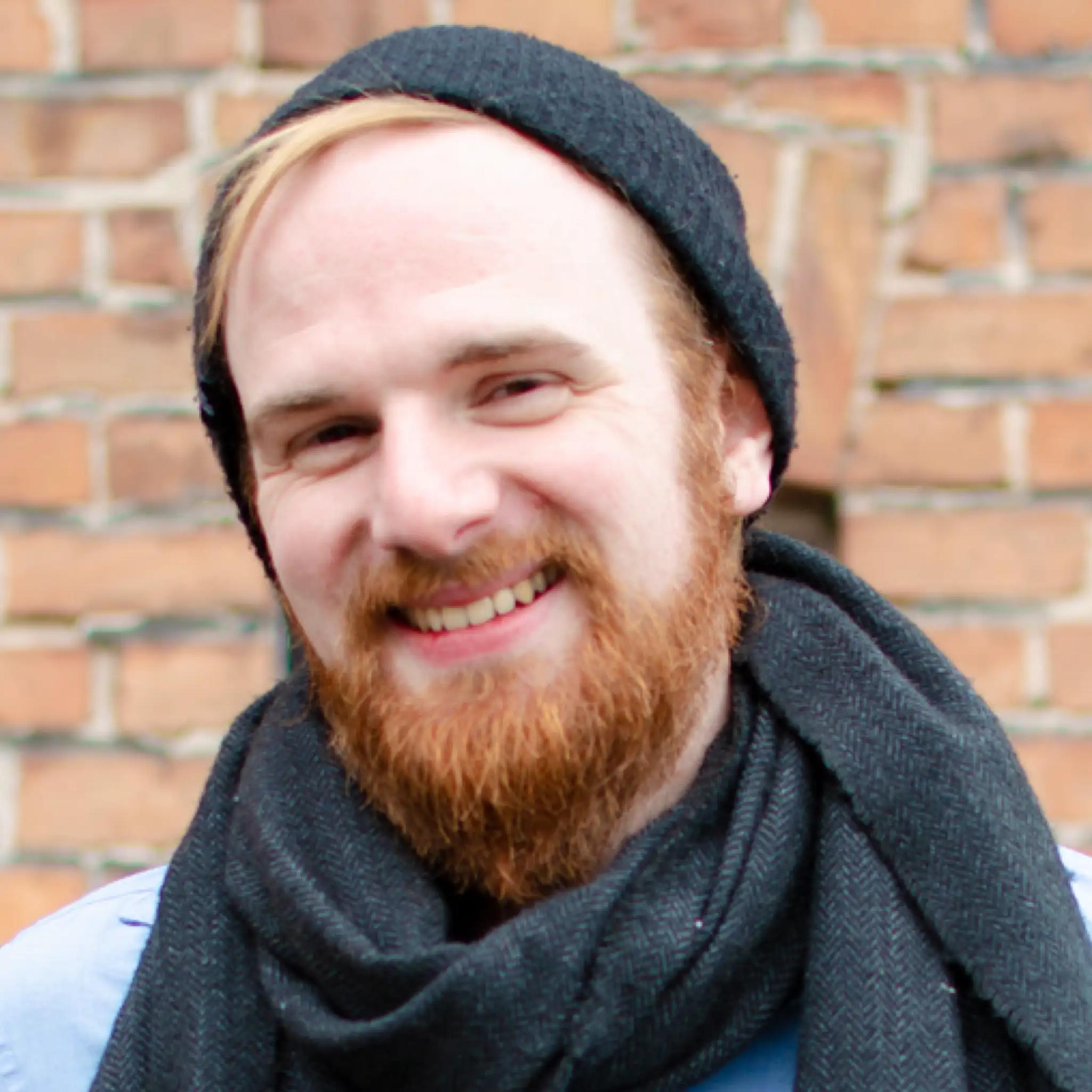 Smiling man with red beard and black beanie in front of brick wall