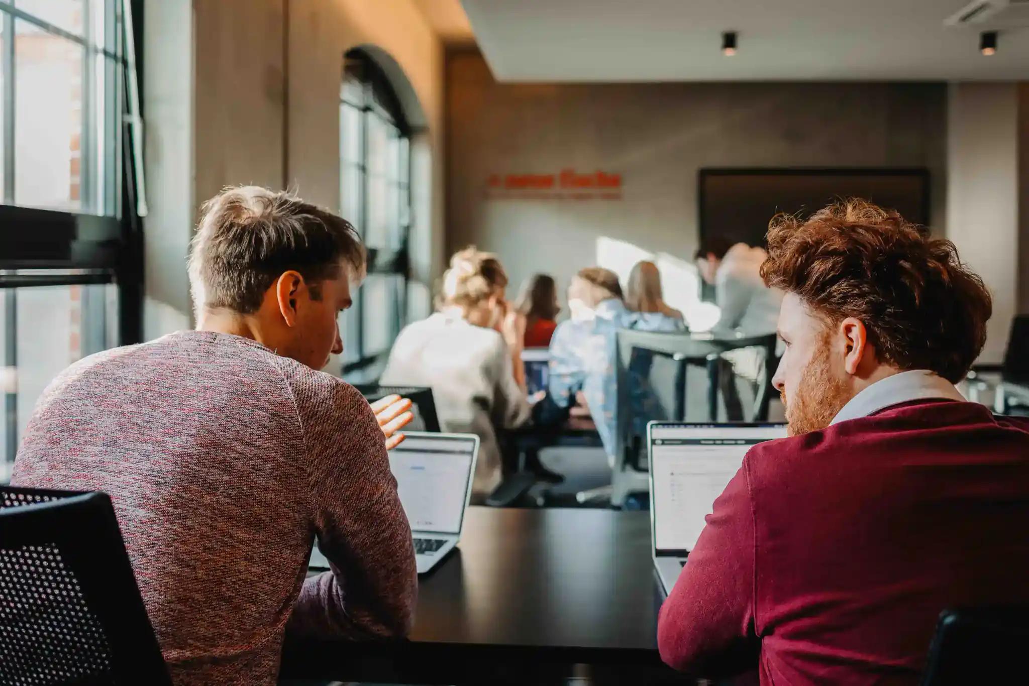 Zwei Männer arbeiten an Laptops in einem Büro mit mehreren Personen.
