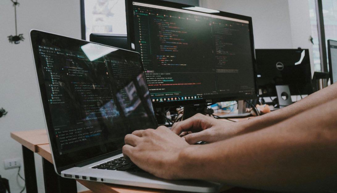 Person tippt Code am Laptop mit Monitor im Hintergrund