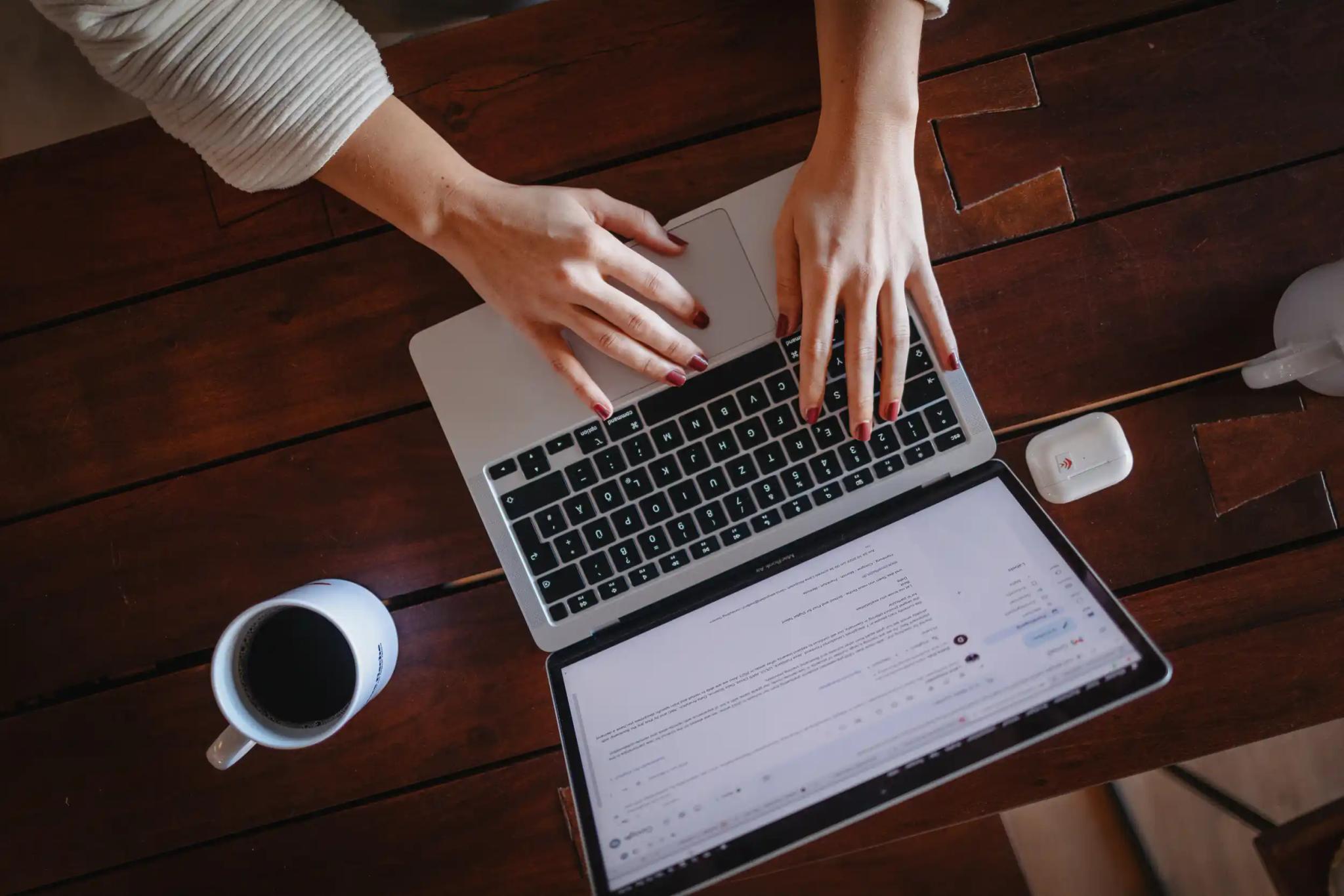 Person typing on laptop keyboard at wooden table with coffee and earphones.