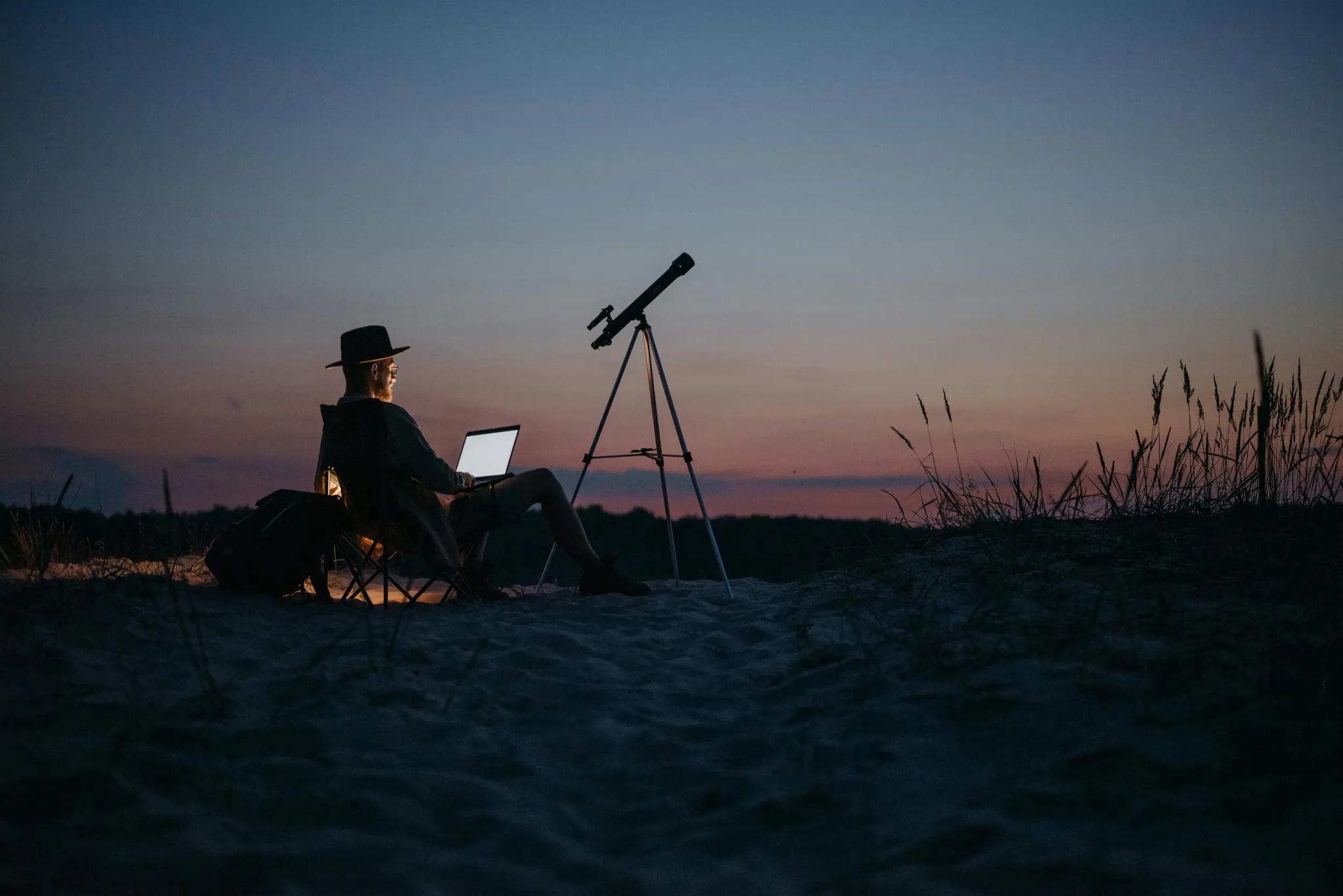 Person with laptop and telescope outdoors at sunset