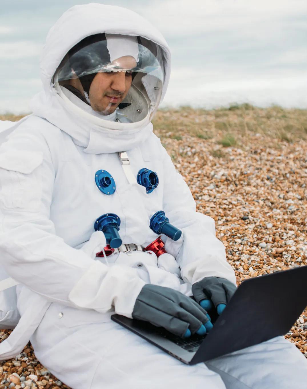 Person im Raumanzug arbeitet am Laptop am Strand
