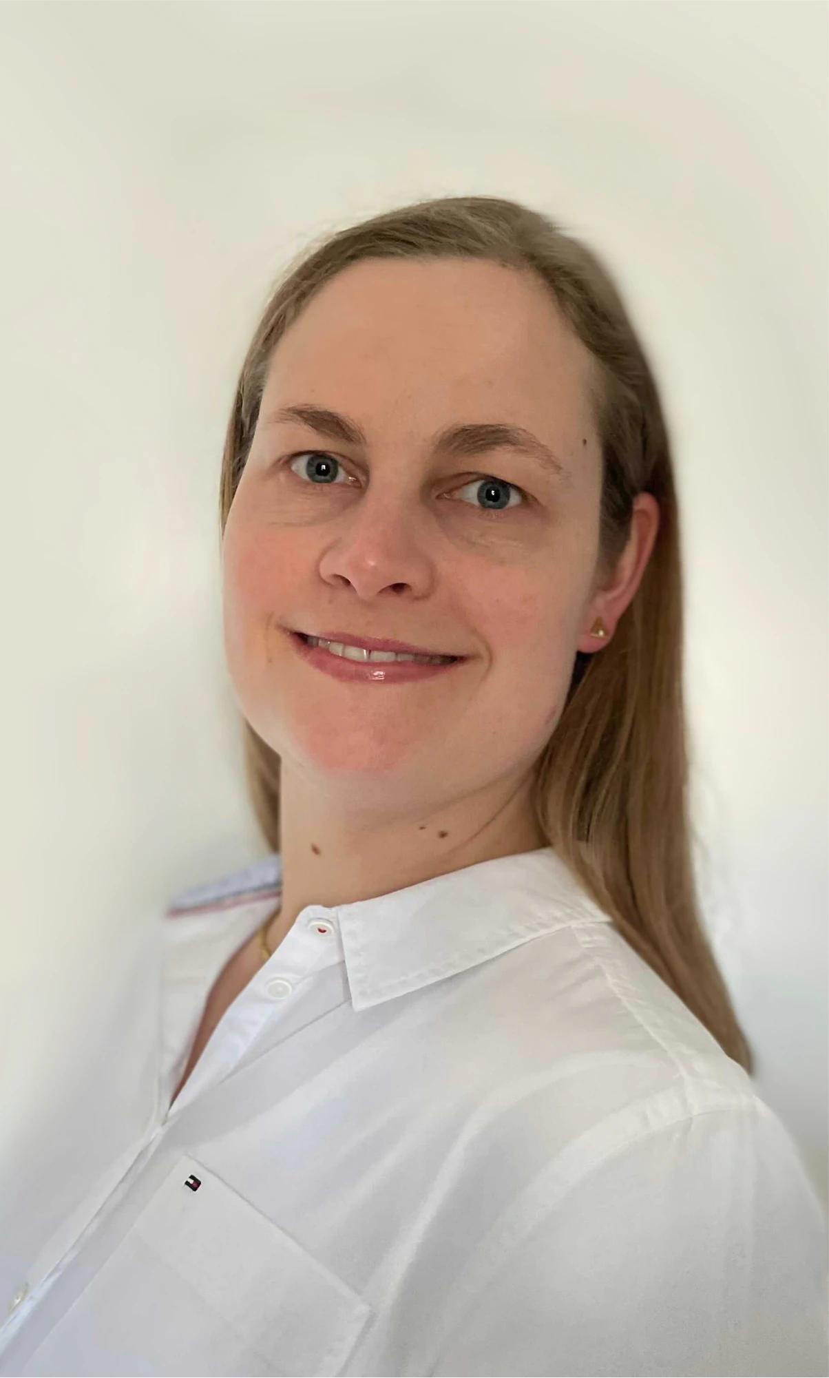 Portrait of a smiling woman with light brown hair and white shirt against a light background.