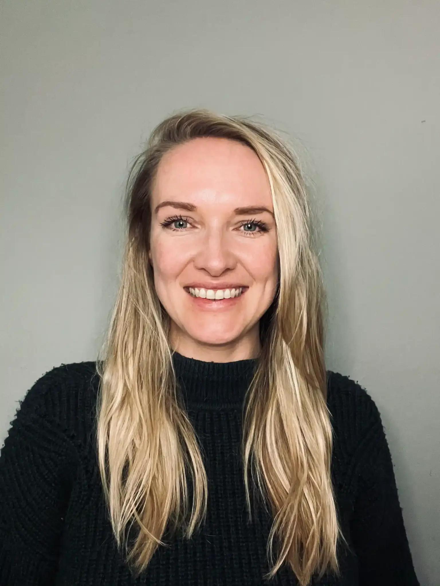 Portrait of a smiling woman with long blonde hair and black sweater against a gray background.