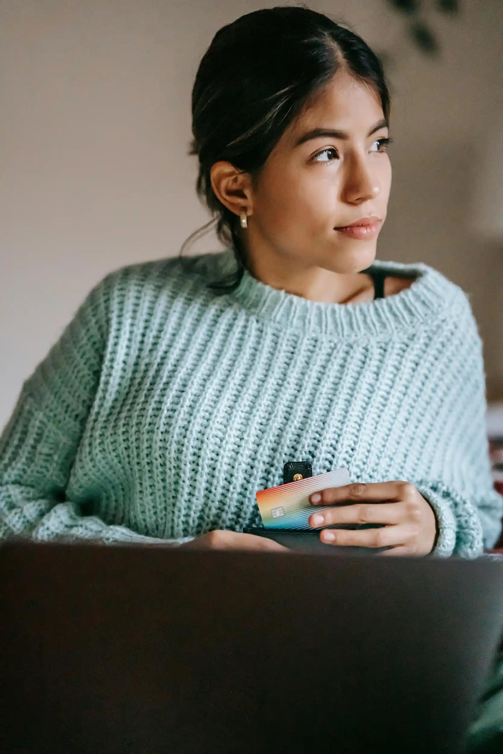 Nachdenkliche Frau in Pullover hält Kreditkarte vor Laptop.