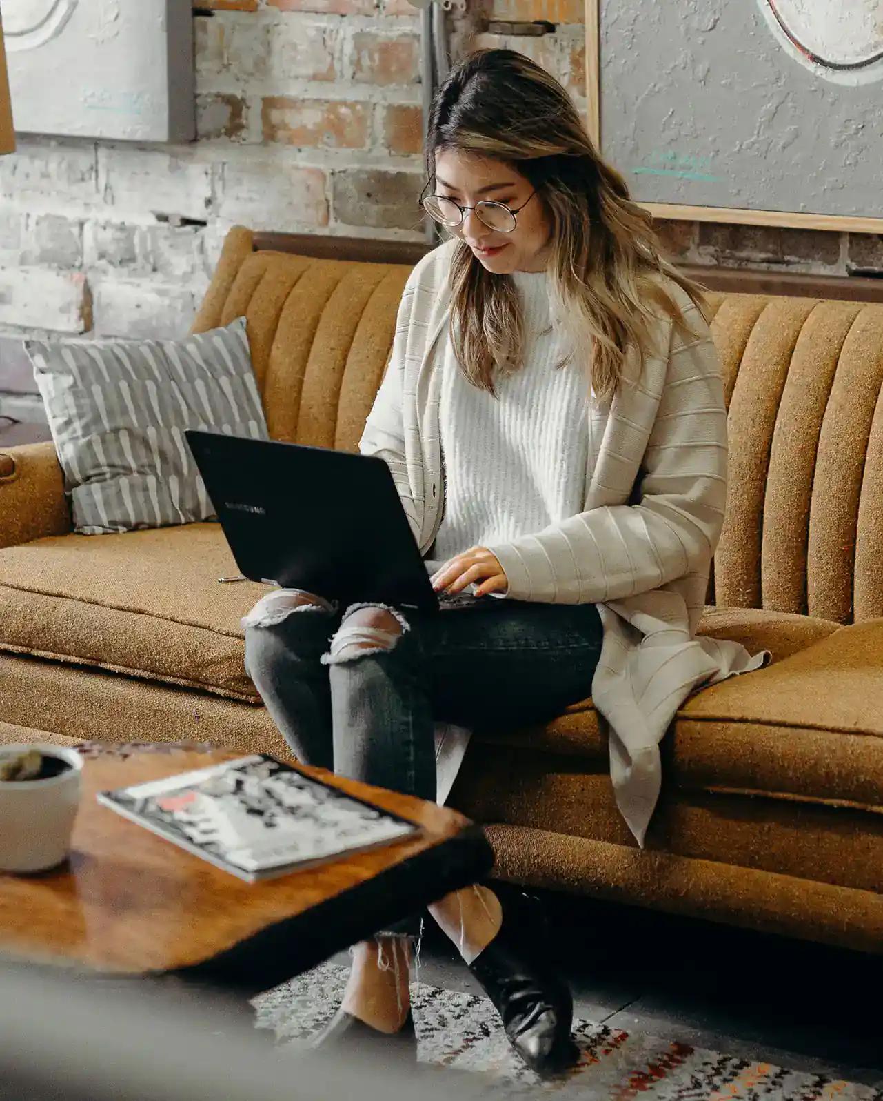 Frau arbeitet mit Laptop auf einer Couch in einem gemütlichen Raum.