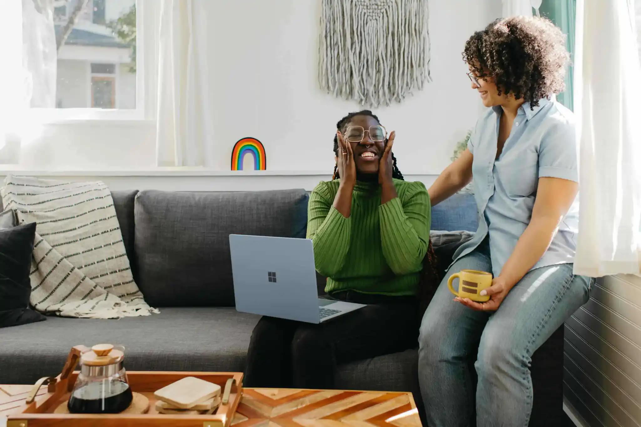 Zwei Frauen lachen zusammen auf einem Sofa mit Laptop.