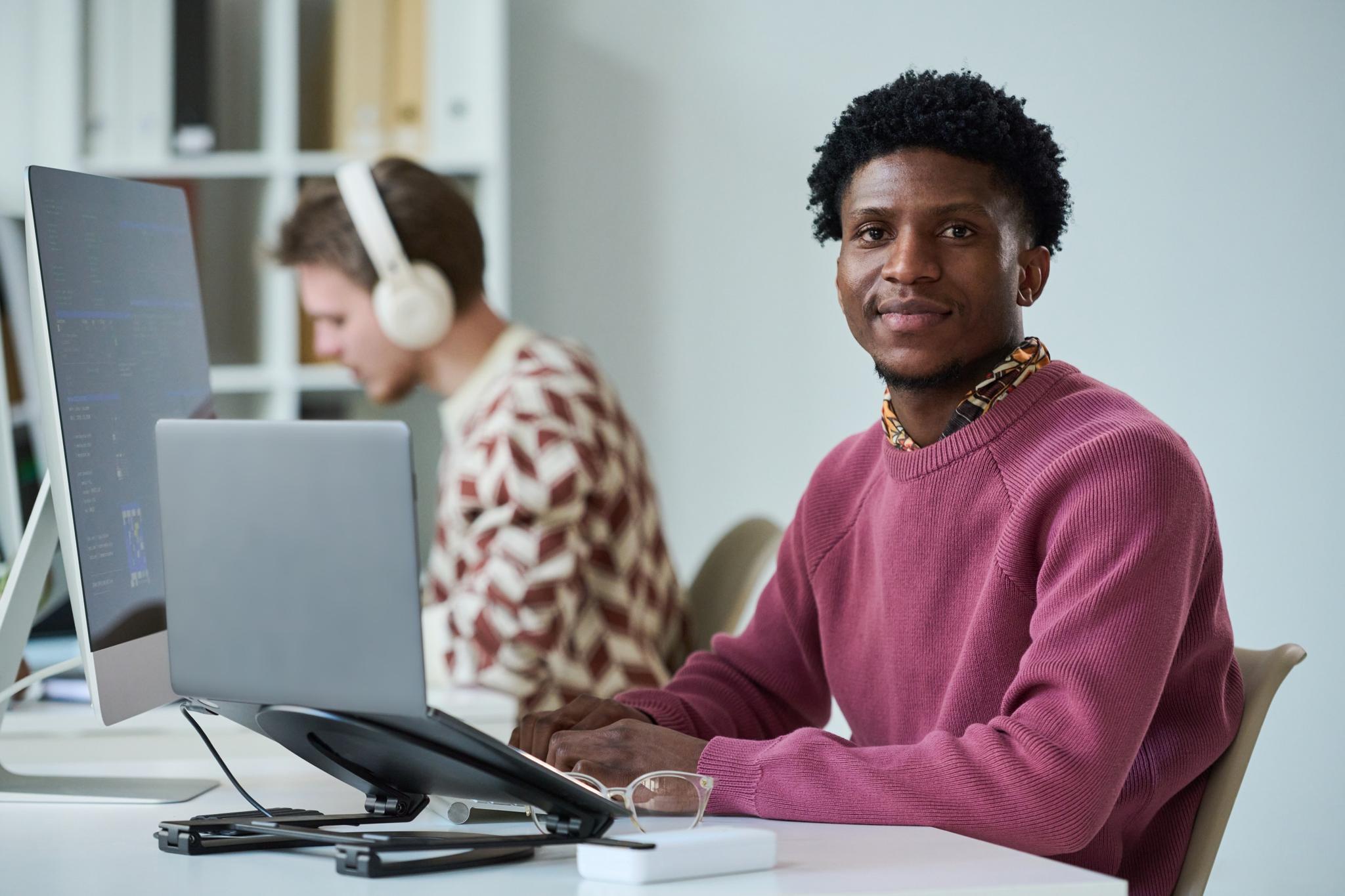 Ein Mann in einem Büro sitzt am Computer, während eine andere Person im Hintergrund Kopfhörer trägt.