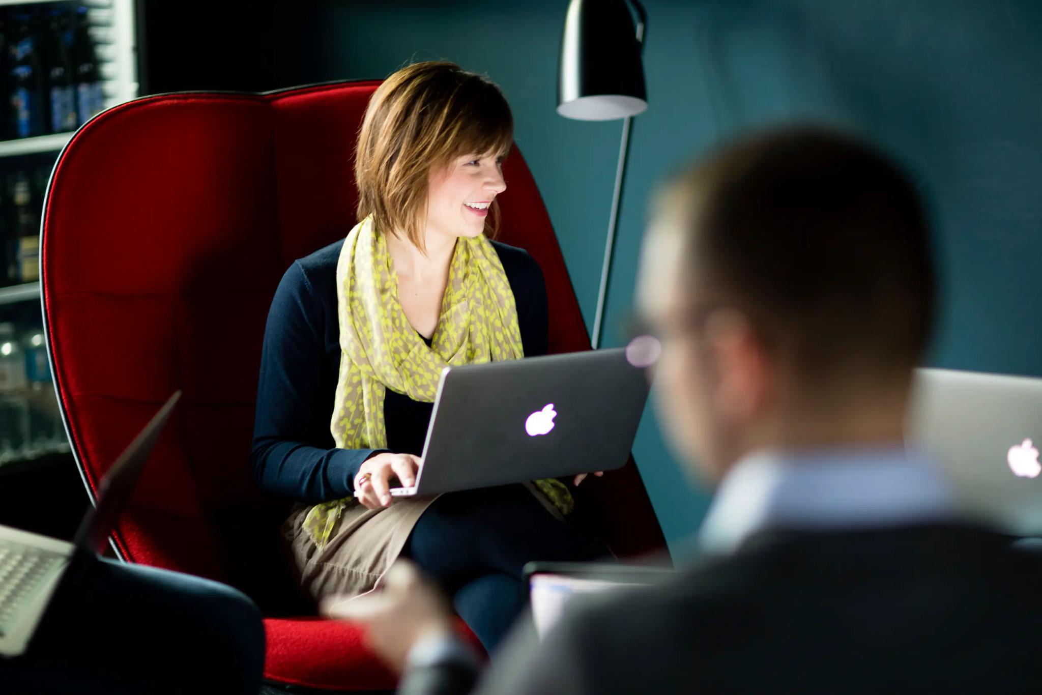 Woman sitting with laptop in red chair talking to colleagues.