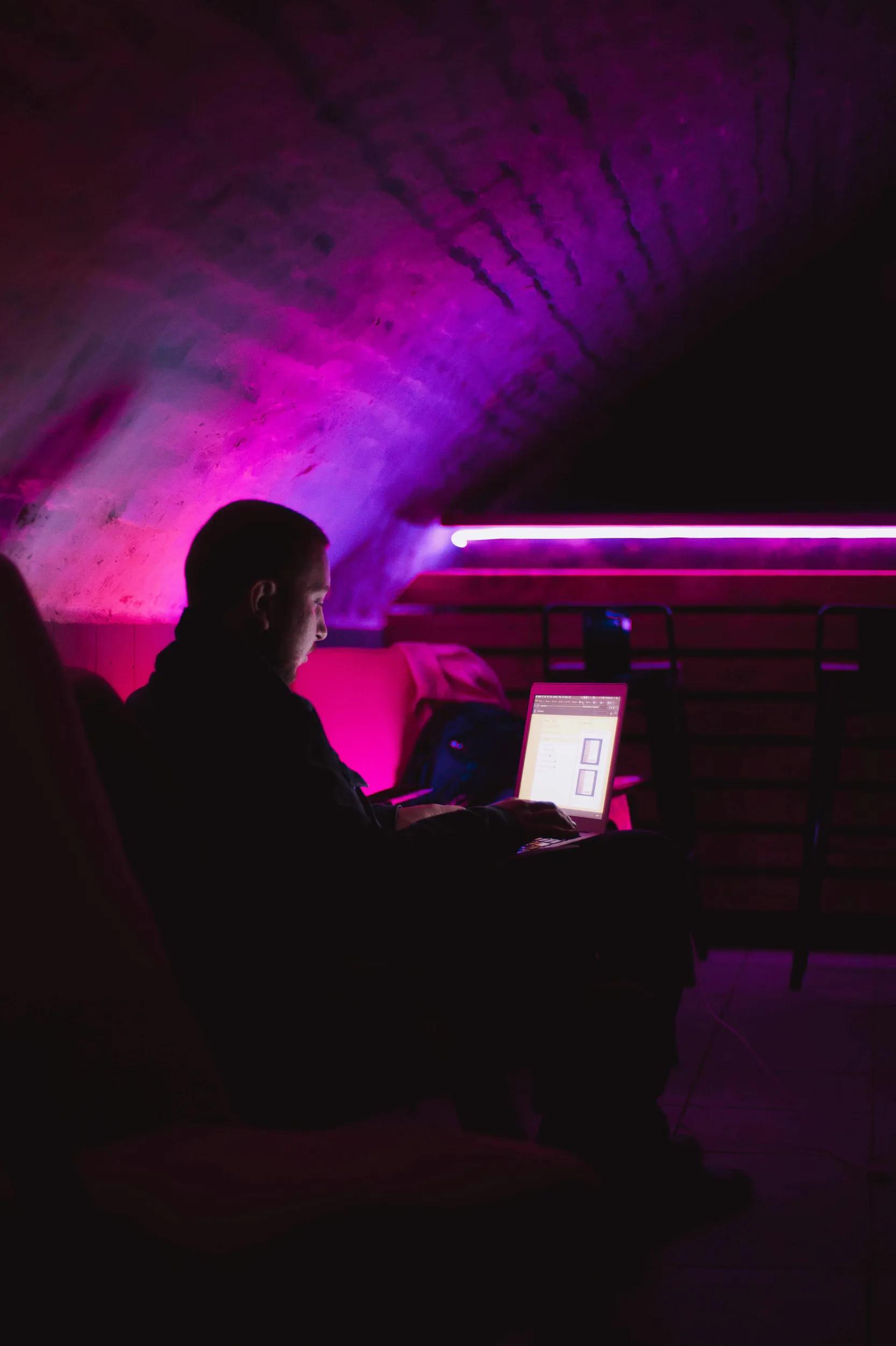 Person sitzt im dunklen Raum mit Laptop und rosa Licht.