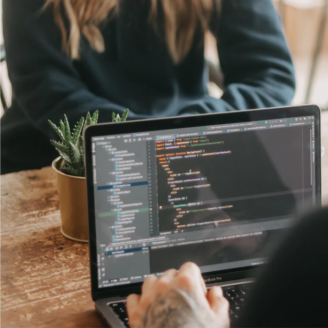Laptop displaying programming code on screen with two people in the background.