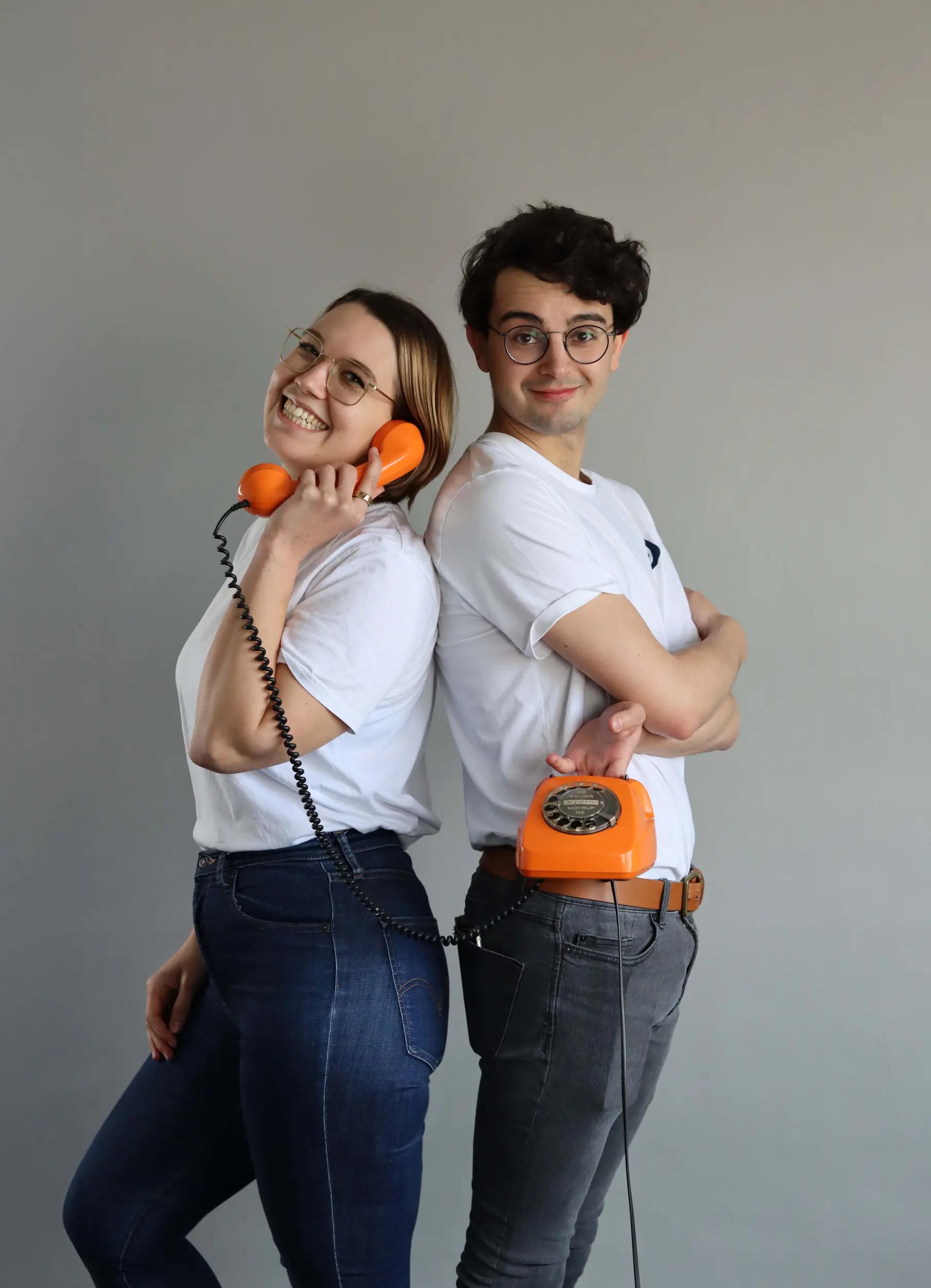 Zwei junge Erwachsene lehnen Rücken an Rücken, eine hält einen orangefarbenen Retro-Telefonhörer, der andere hält die Basis des Telefons.